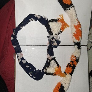 May include: A headband featuring a dark blue fabric with a white floral pattern and two attached crocheted ears, accompanied by a long, segmented crocheted tail. The ears and tail are crafted in a calico pattern of white, orange, and black yarn.