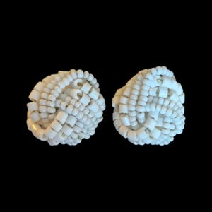 May include: Two white beaded earrings. Each earring is a rounded shape, constructed from small, square-shaped beads. The beads are tightly woven together, creating a textured surface. The earrings are set against a black background.