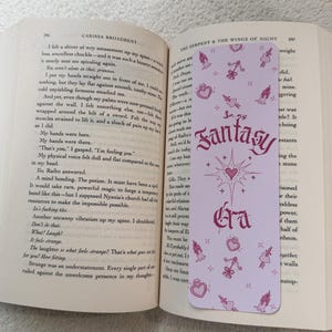 May include: A light purple bookmark with the text "In my Fantasy Era" in a decorative font. The bookmark features pink illustrations of hearts, bows, and potion bottles. The bookmark is placed in an open book.