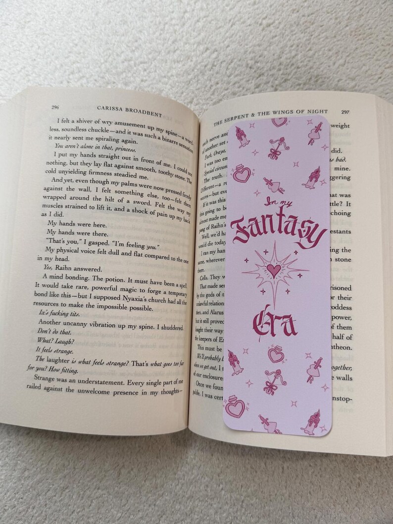 May include: A light purple bookmark with the text "In my Fantasy Era" in a decorative font. The bookmark features pink illustrations of hearts, bows, and potion bottles. The bookmark is placed in an open book.