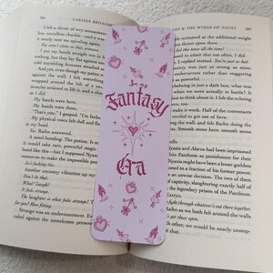 May include: A light purple bookmark with the text "In my Fantasy Era" in a decorative font. The bookmark features small pink illustrations of hearts, perfume bottles, and figures. The bookmark is placed in an open book.