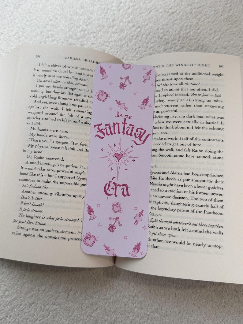 May include: A light purple bookmark with the text "In my Fantasy Era" in a decorative font. The bookmark features small pink illustrations of hearts, perfume bottles, and figures. The bookmark is placed in an open book.