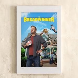 Op de afbeelding: Een ingelijste filmposter voor The Breadwinner met een persoon die een witte mok vasthoudt met de tekst #2 PARENT. De achtergrond toont een huis in aanbouw met steigers, een paard en andere figuren bij de veranda.