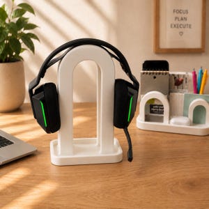 May include: A white arch-shaped headphone stand holding black gaming headphones with green LED accents. Beside it sits a white desk organizer containing a memo book, pens, and small items, with a framed sign reading FOCUS PLAN EXECUTE.