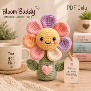 May include: A crochet flower toy with a yellow face, black eyes, a smiling mouth, and pink cheeks. The petals are purple, pink, and orange. The stem is green with two leaves and a pink heart. A tag reads: You are growing beautifully.