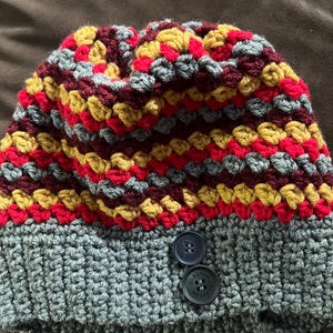 Puede incluir: Un gorro de ganchillo hecho a mano con un borde gris y una parte superior colorida. La sección superior presenta un patrón de hilo rojo, amarillo, gris y burdeos. Dos botones azul oscuro están cosidos en el borde. El gorro es un accesorio acogedor.