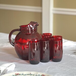 May include: A set of deep red glass drinkware consisting of one large rounded pitcher with a curved handle and spout, accompanied by several matching tall, cylindrical drinking glasses, all featuring a smooth, translucent finish.