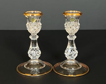 Puede incluir: Un par de candelabros de cristal transparente con un patrón de corte de diamante en los tallos y las arandelas. Cada candelabro tiene una abertura superior con borde dorado y una base circular dorada, con una pequeña pegatina visible.