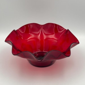 Könnte beinhalten: Eine leuchtend rote Glasschale mit gewelltem Rand und dunklem Rand. Die Schale hat eine breite, offene Form und steht auf einem kleinen, kreisförmigen Sockel. Das Glas hat eine glänzende Oberfläche.