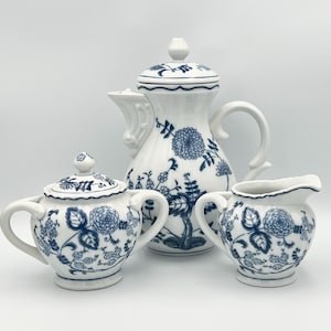 Może przedstawiać: Biały porcelanowy zestaw do herbaty z niebieskimi kwiatowymi wzorami. Zestaw zawiera czajniczek, cukiernicę z pokrywką i dzbanuszek do śmietanki. Czajniczek ma wysoki, zakrzywiony dzióbek i zaokrąglony korpus.
