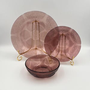 May include: A set of three pieces of vintage glassware in shades of purple. The set includes two plates and a bowl. The plates are displayed on gold-colored stands. The glassware has a textured, radial design.