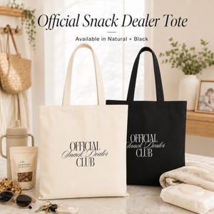 May include: Two canvas tote bags, one natural beige and one black, each featuring the text "OFFICIAL Snack Dealer CLUB" printed in a serif font. The bags have two shoulder straps and a rectangular shape, displayed side by side on a flat surface.