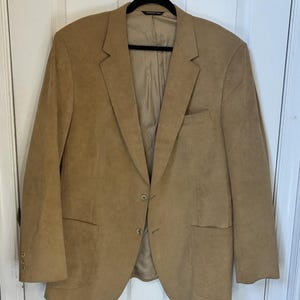 May include: A tan blazer with a two-button closure, notched lapels, and two front pockets. The blazer is hanging on a black hanger. The blazer is made of a suede-like material.