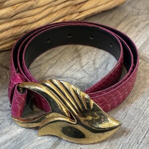 Peut inclure: Une ceinture magenta avec un motif texturé en peau de serpent et une grande boucle en métal doré poli en forme de feuille ou d'aile stylisée. La ceinture présente une doublure intérieure noire et plusieurs trous de réglage sur la sangle.