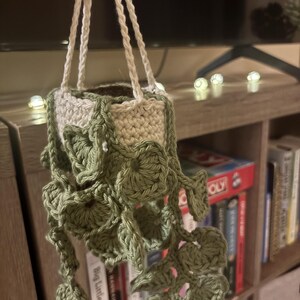 May include: A crocheted hanging plant in a cream-colored pot with long, trailing vines featuring multiple sage green leaves. The plant is suspended by three thin, cream-colored crocheted cords that meet at the top for hanging display.