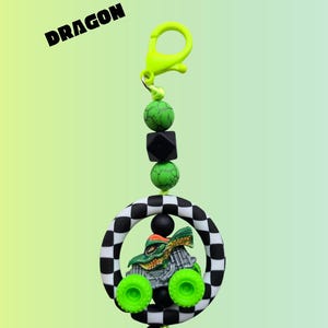 K&ouml;nnte beinhalten: Ein gr&uuml;nes und silbernes Monstertruck-Spielzeug mit neongr&uuml;nen R&auml;dern h&auml;ngt in einem schwarz-wei&szlig; karierten Ring. Der Schl&uuml;sselanh&auml;nger hat einen neongr&uuml;nen Verschluss und ist mit gr&uuml;nen und schwarzen Perlen verziert. Das Wort "DRAGON" ist schwarz gedruckt.