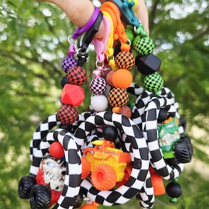 K&ouml;nnte beinhalten: Ein farbenfroher Schl&uuml;sselanh&auml;nger mit einer Vielzahl von Perlen, darunter karierte, orangefarbene und schwarze Kugeln sowie ein Monstertruck-Spielzeug. Der Schl&uuml;sselanh&auml;nger hat eine Mischung aus Farben, darunter Rot, Orange, Gr&uuml;n und Schwarz, und wird von farbigen Verschl&uuml;ssen zusammengehalten.