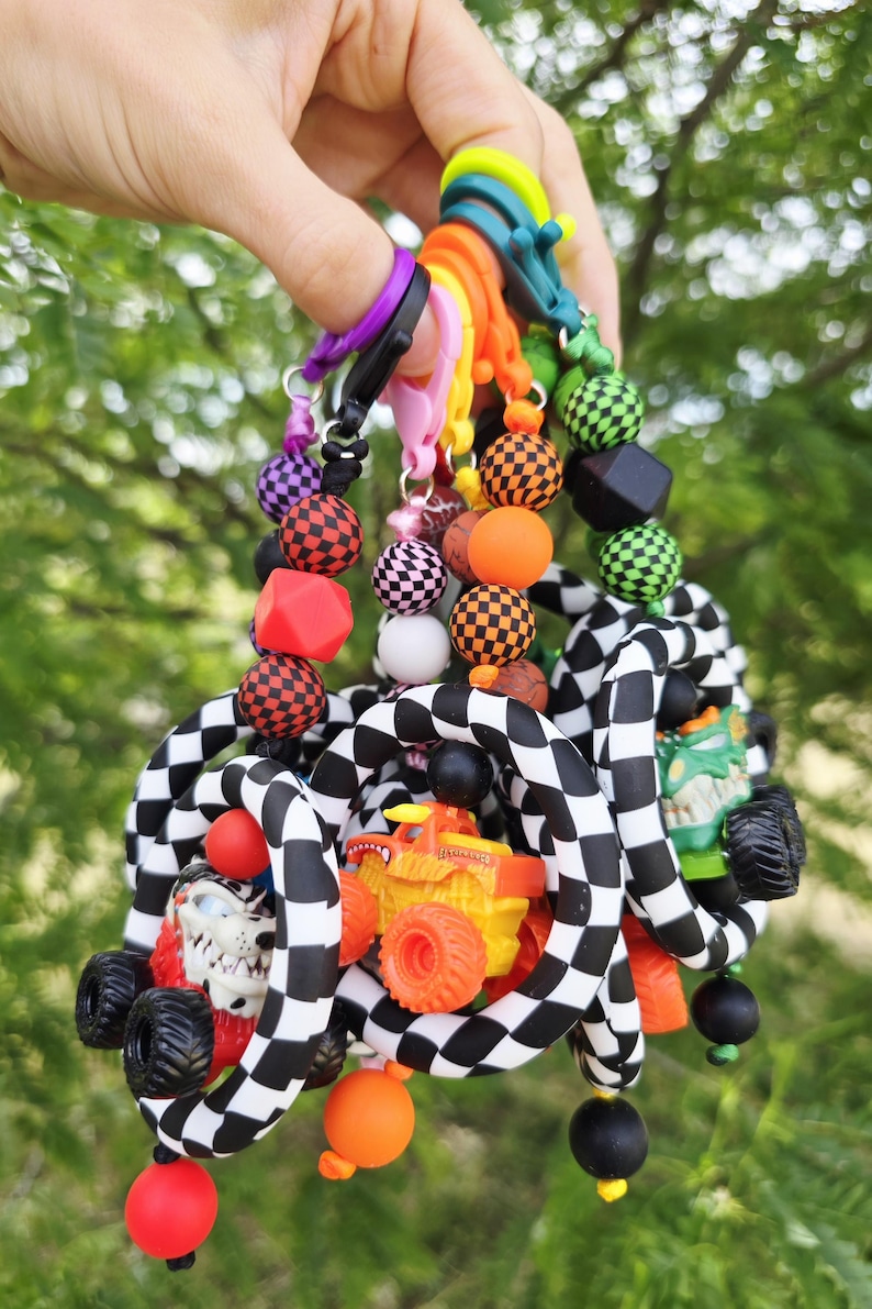 K&ouml;nnte beinhalten: Ein farbenfroher Schl&uuml;sselanh&auml;nger mit einer Vielzahl von Perlen, darunter karierte, orangefarbene und schwarze Kugeln sowie ein Monstertruck-Spielzeug. Der Schl&uuml;sselanh&auml;nger hat eine Mischung aus Farben, darunter Rot, Orange, Gr&uuml;n und Schwarz, und wird von farbigen Verschl&uuml;ssen zusammengehalten.