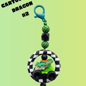 K&ouml;nnte beinhalten: Ein skurriler Schl&uuml;sselanh&auml;nger mit einem Cartoon-Drachen in einem Monstertruck. Der Drache ist gr&uuml;n mit goldenen Akzenten, eingebettet in einen schwarz-wei&szlig; karierten Ring. Der Text "Cartoonized Dragon #2" ist sichtbar.