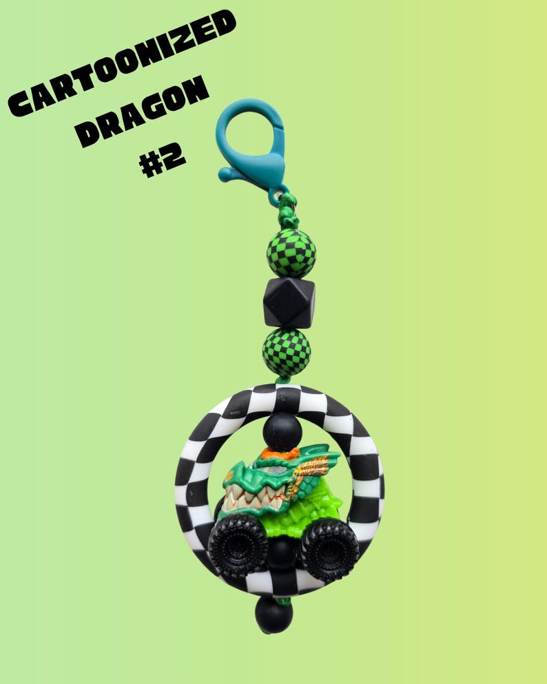 K&ouml;nnte beinhalten: Ein skurriler Schl&uuml;sselanh&auml;nger mit einem Cartoon-Drachen in einem Monstertruck. Der Drache ist gr&uuml;n mit goldenen Akzenten, eingebettet in einen schwarz-wei&szlig; karierten Ring. Der Text "Cartoonized Dragon #2" ist sichtbar.