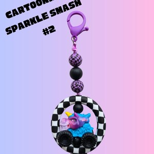 K&ouml;nnte beinhalten: Ein Cartoonized Sparkle Smash Schl&uuml;sselanh&auml;nger mit einem lila Clip, schwarzen und lila Perlen und einem Monstertruck mit einem Einhorn-Design in einem schwarz-wei&szlig; karierten Kreis. Unten befindet sich eine schwarz-wei&szlig;e Schleife.