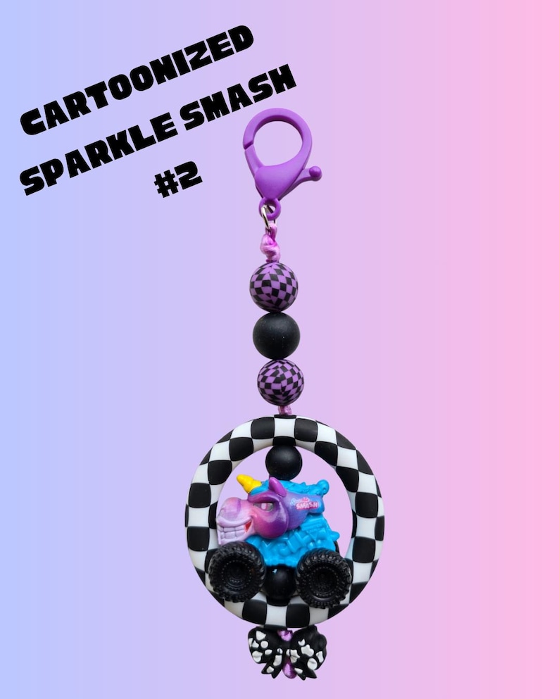 K&ouml;nnte beinhalten: Ein Cartoonized Sparkle Smash Schl&uuml;sselanh&auml;nger mit einem lila Clip, schwarzen und lila Perlen und einem Monstertruck mit einem Einhorn-Design in einem schwarz-wei&szlig; karierten Kreis. Unten befindet sich eine schwarz-wei&szlig;e Schleife.