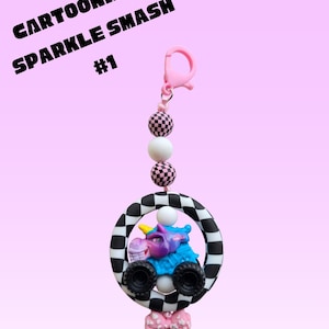K&ouml;nnte beinhalten: Ein Cartoonized Sparkle Smash Schl&uuml;sselanh&auml;nger mit einem rosa Verschluss, karierten Perlen und einem Cartoon-Monster-Truck in einem schwarz-wei&szlig; karierten Kreis. Unten befindet sich eine rosa Schleife. Der Text "Cartoonized Sparkle Smash #1" steht oben.