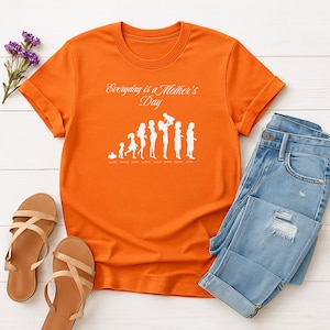 May include: Orange t-shirt with the text "Everyday is a Mother's Day" and a white graphic of a mother and child through the years. The shirt is paired with blue jeans and brown sandals.