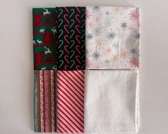 Può includere: Un set di sei quadrati di tessuto piegati con vari motivi natalizi, tra cui verde con alberi rossi e renne bianche, nero con bastoncini di zucchero rossi, bianco con fiocchi di neve colorati, bordi a righe, strisce diagonali rosse e bianche e bianco tinta unita.