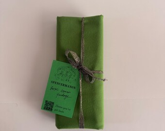 Può includere: Un pezzo di tessuto verde tinta unita piegato, legato con un sottile nastro metallico argentato. È attaccata un'etichetta verde rettangolare con il testo SPENCERMANON, Basic Green Yardage e Damn you've got taste, insieme a un codice QR.