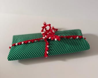 Può includere: Un articolo in tessuto rettangolare con sfondo verde e motivo a pois bianchi. L'articolo è avvolto da un nastro rosso con piccoli pon-pon bianchi lungo tutta la lunghezza, annodato con un fiocco nella parte superiore centrale.