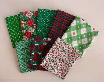 Può includere: Una collezione di otto quadrati di tessuto piegati con vari motivi natalizi in rosso, verde e bianco. I disegni includono tartan, vichy, renne, pini, foglie di agrifoglio e forme geometriche disposte a ventaglio.