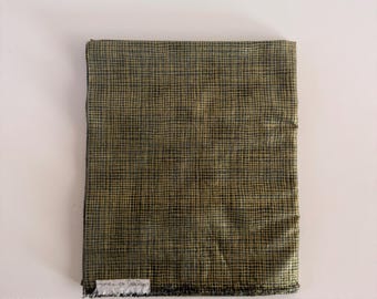 Può includere: Un pezzo di tessuto rettangolare piegato con un motivo a griglia incrociata verde oliva scuro e nero. Una piccola etichetta in tessuto bianco con testo nero è attaccata all'angolo inferiore sinistro del tessuto, che presenta un bordo leggermente sfilacciato.