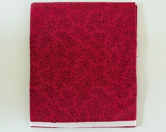 Può includere: Un pezzo di tessuto rosso piegato con un motivo botanico ripetitivo di piccole foglie e steli sottili in marrone scuro o nero. Il tessuto presenta una cimosa bianca pulita e dritta visibile lungo il bordo inferiore della piega.