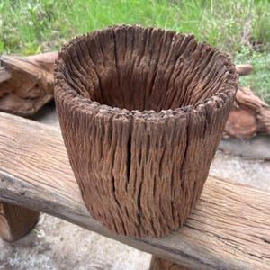 Könnte beinhalten: Ein zylindrischer Pflanzkübel aus einem ausgehöhlten Stück verwittertem braunem Holz. Die Außen- und Innenflächen weisen eine tief gerillte, vertikale Maserung auf, die das natürliche, raue und gealterte Erscheinungsbild des Holzes unterstreicht.