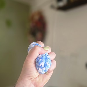 May include: A small, round, plush object crafted from soft, variegated yarn in shades of light blue, white, and dark blue. The texture is fuzzy and tactile, featuring a dense, looped knit construction that is held firmly in a closed fist.
