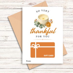 Può includere: Un biglietto d'auguri di 13x18 cm con busta color sabbia, decorato con un'illustrazione di una candela accesa, eucalipto e una fetta d'arancia essiccata. Il testo recita SO VERY thankful FOR YOU e GIFT CARD. Una carta regalo arancione con fiocco.