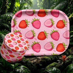 May include: A pink rolling tray and a matching round container, both adorned with a repeating pattern of red and pink strawberries. The tray has rounded edges. The background is a lush green jungle.