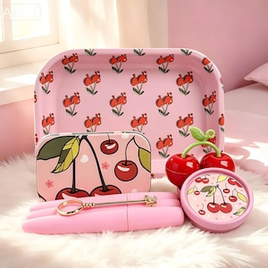 May include: A collection of pink cherry-themed accessories, including a rectangular tray, a rectangular tin, a small round container, and a pink pen holder with a gold-colored pen. The tray and tin feature a repeating cherry pattern.