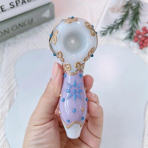 May include: A decorative glass pipe with a light purple stem and a white bowl. The stem features blue star and dot designs, with gold accents. The bowl has gold swirls and blue accents. The pipe is held in a hand.