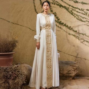 Puede incluir: Un vestido largo color crema con mangas largas y falda hasta el suelo. El vestido presenta botones y adornos dorados en la parte delantera y alrededor de la cintura. Las mangas tienen un ligero abullonado en la muñeca. El vestido es de tela suave y fluida.