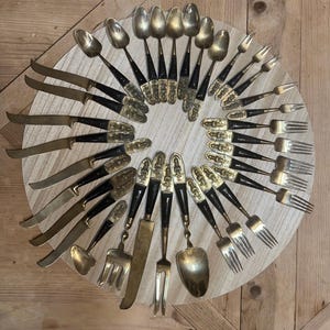 May include: A collection of gold-toned metal flatware arranged in a circular pattern. The set includes various knives, spoons, and forks, each featuring black handles and ornate, embossed gold-colored decorative accents at the top of the stems.