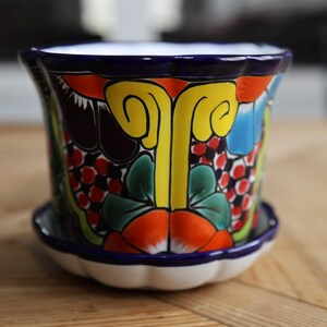 May include: A hand-painted ceramic flower pot with a matching saucer. The pot features a vibrant floral design in yellow, orange, red, blue, and green against a dark blue background. The rim and saucer edge are also dark blue.