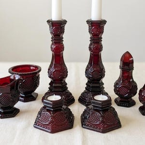May include: A collection of deep red pressed glass items featuring a repeating diamond and circle pattern. The set includes two tall candlesticks with white candles, two votive candle holders, a small creamer pitcher, a sugar bowl, and two shakers.