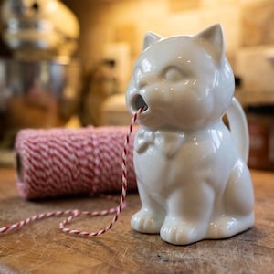 May include: A white ceramic cat-shaped creamer with a bow tie, holding a red and white twine in its mouth. A roll of red and white twine sits behind the creamer on a wooden surface. The creamer has a handle on its side.