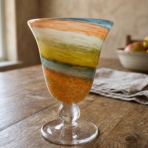 May include: A glass goblet featuring a flared bowl with horizontal bands of orange, white, yellow, and blue colors. The stem includes a clear glass sphere above a circular base, providing a stable foundation for the colorful, textured glass vessel.