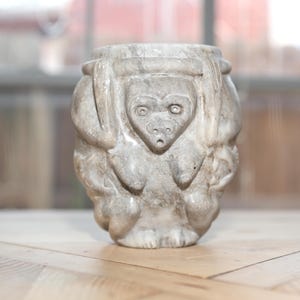 May include: A carved stone vessel with a monkey face design. The gray and white sculpture has a cylindrical shape with a detailed monkey face in the center and other monkey features around the sides. The vessel is likely for decorative purposes.