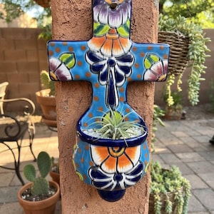 May include: A wall-mounted ceramic planter shaped like a cross, featuring a vibrant blue base with hand-painted floral patterns in orange, purple, and white. The lower section forms a rounded basin containing a small air plant nestled in white gravel.
