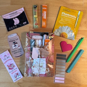 May include: An assortment of self-care and reading-themed items on a wooden surface. Includes a rose eye mask, tea and coffee packets, a chamomile face mask, a heart-shaped item, pens, sticky notes, and a gift bag with floral print.