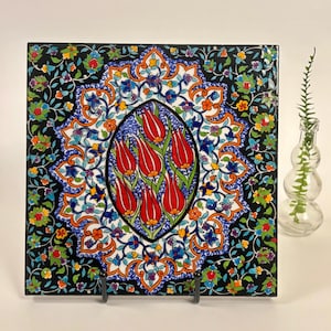 May include: A square ceramic tile featuring a central oval medallion filled with six red tulips on green stems. The surrounding border displays a dense pattern of colorful flowers and vines against a black background, finished with a glossy glaze.